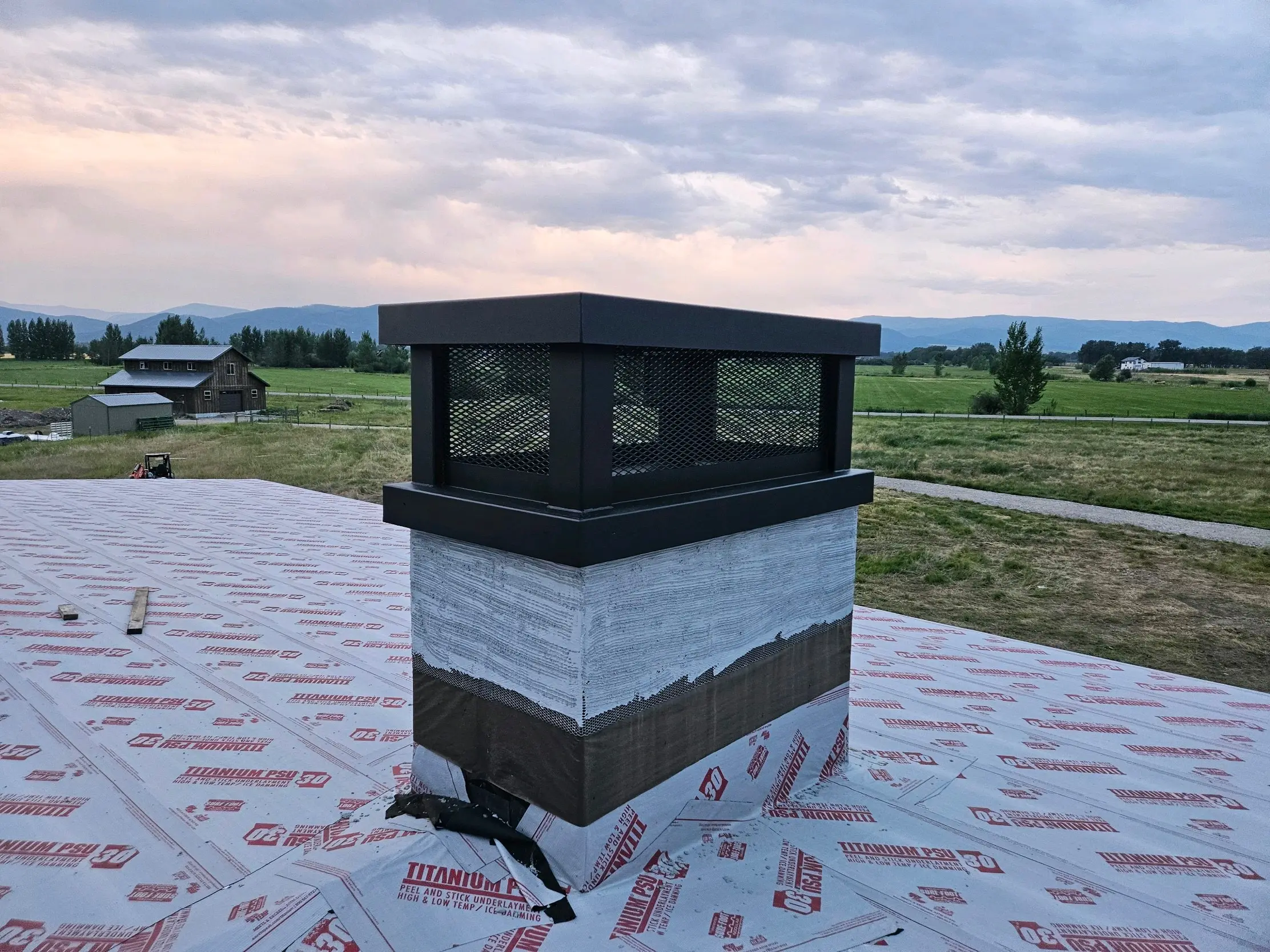A black steel chimney cap with mesh sides on a roof at dusk, with the Gallatin Valley beyond