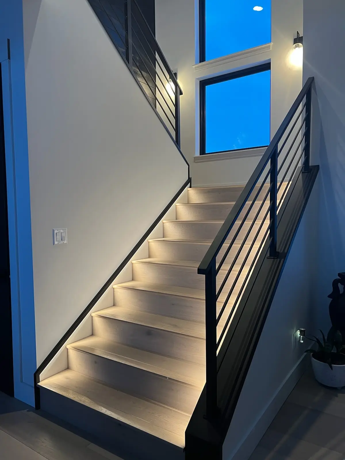 A staircase at night with light oak treads, a black horizontal-bar railing, and lit steps